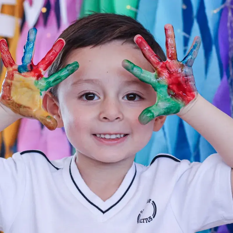 Niño sonriente con manos pintadas de colores frente a fondo arcoíris durante actividad artística escolar